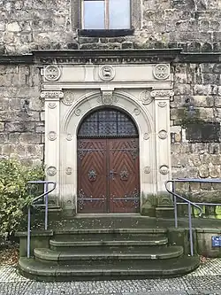 Uprostřed fotografie hnědě natřené dveře, okolo nich světle edý pískovcový portál s mnoha ozdobami, v pozadí tmavá stěna kostela, vepředu schodiště