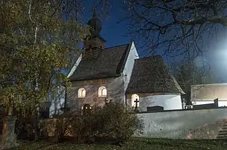 Fotografie jižní stěny kostela s presbytářem v noci, před kostelem kamenná zeď, vlevo strom a kříž