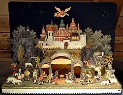 Příbramské jesličky se sbírky Sieglinde a Udo Hergesell, vpravo mužská postava pastýře v červeném kabátci daruje vánočku