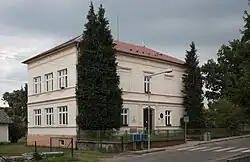 Patrová zděná, světle omítnutá budova školy schovaná za třemi vzrostlými jehličnatými stromy mírně svoji výškou přesahujícími budovu samotnou