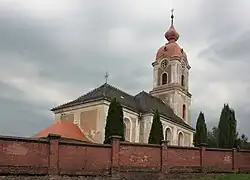 Zděná sakrální stavba, před níž je patrná hřbitovní zeď, vybudovaná z červených cihel