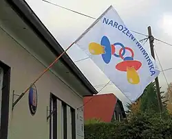 Vlajka visící z domu. Vlajka má bílé pozadí a na něm je obraz dvou spojených dudlíků, nad nimiž je text „Narození miminka“.