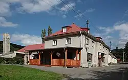 Sluncem osvícený patrový zděný objekt restaurace. V popředí stojí dřevěná přístavba zastřešené letní zahrádky. Vpravo kolem budovy restaurace prochází zpevněná silnice.