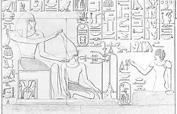 Kopie reliéfu. Na levé straně na trůnu sedí polonahý muž s přikrývkou hlavy, sukní a bradkou, který pohlíží na pravou stranu. Napravo zády k němu klečí na pravou stranu hledící muž, jenž má na hlavě korunu, které se ze shora svojí levou rukou dotýká muž nalevo. Klečící muž je polonahý, má bradku a sukni, ruce má svěšené dolů, jeho krku se dotýká sedící muž napravo svojí pravou rukou. Na druhé straně výjevu na proti klečícímu muži nalevo klečí další muž se šaty, pokrývkou hlavy a bradkou, levou ruku má pozvednutou nalevo a pravou ruku má svěšenou dolů. Kolem celého výjevu se nacházejí hieroglyfy a symboly.