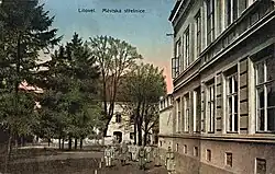 Kolorovaná pohlednice s pohledem na budovu střelnice. Budova se nachází vzadu za stromy a v popředí stojí vojáci v uniformách. Zcela napravo v popředí je zčásti vidět další vysoká budova. Pod horním okrajem pohlednice je nápis „Litovel. Městská střelnice.“