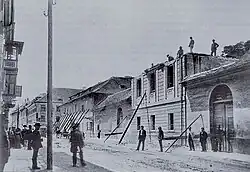 Černobílá fotografie domů poničených zemětřesením, jejich zbytky jsou podepřené kůly, na ulici a na střechách osoby