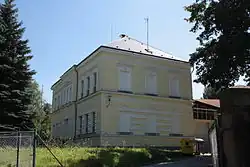 Boční pohled na objekt bývalé školy. Její patrová žlutě zbarvená budova stojí na mírném vršíčku. V popředí je veden pletivový plot.