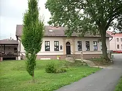 Ústřední motiv tvoří jednopatrová budova bývalé školy, která složí jako obecní úřad. Na budově u dveří jsou dva nápisy obecní úřad a místní veřejná knihovna. Nalevo se nachází dřevěná zahrádka místního hostince. Napravo vzadu je vidět část budovy bývalého obchodu. Na trávníku před budovou obecního úřadu rostu keře a jeden vzrostlý strom. Pravým spodním okrajem vede místní komunikace a od ní se odděluje schodiště směřující do budovy úřadu.