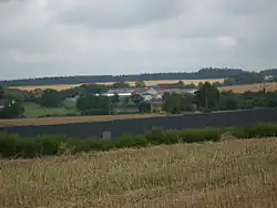 Vepředu se nachází pole s posekaným strništěm. Uprostřed stojí tmavěšedá protihluková stěna u dálnice D1, za ní je vidět posečené pole a louka a řada vzrostlých stromů. Za nimi stojí čtyři budovy zemědělského družstva, z nichž je jedna s červenou střechu. Za budovami se nachází další pole a úplně vzadu obrysy lesa.