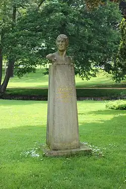 Pískovcová busty na soklu ze stejného materiálu umístěná v travnaté ploše.