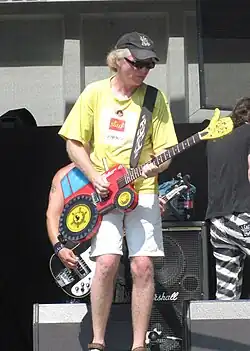 Záběr na osluněného Ivana Ruta na pódiu, se žlutým tričkem alba Visacího zámku 02 Start, černou kšiltovkou Visací zámek a šedými kraťasy; v ruce drží kytaru s tělem ve tvaru červeného traktoru Zetor se žlutými koly a s hlavou ve tvaru zdviženého palce.