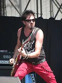 Drsně se tvářící Michal Pixa s černými brýlemi oděný v červených kraťasech a černém tílku Visací zámek na pódiu při hraní našlapuje energicky na pravou nohu.