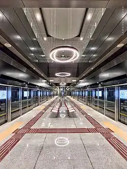 Pohled na nástupiště podzemní stanice metra. Po obou stranách jsou prosklené posuvné dveře nástupiště. Interiér působí moderně, uprostřed nástupiště jsou sedačky. Podlaha z dlaždic šedé a hnědé barvy, strop je vysoký.