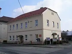 Dvoupodlažní budova bývalé školy v těsném sousedství kostela svatého Jiří. Budova školy je zakončena mansardovou střechou. Před budovou školy se nachází asfaltová plocha křižovatky. Vpravo od budovy školy do oblouku stojí tři vitríny.