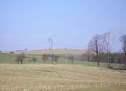 V popředí se nachází posekané pole, jež je vzadu ohraničeno vzrostlými stromy od louky. V pozadí stoupá Mořkovský vrch, na kterém ve základnová stanice mobilního operátora a stožáry vysokého elektrického napětí.