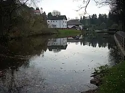 Ve vodní ploše rybníka se odráží okolní domy, na pravé straně část zábradlí, vlevo zeleň