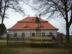 V pozadí stojí farní budova s červenou mansardovu střechou, vepředu oplocená socha svatého Jana Nepomuckého bez koruny, před plotem lavička