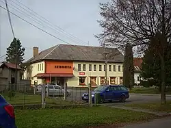 Žlutooranžová budova s eternitovou střechou, vepředu parkující auta a roh oplocené farské zahrady