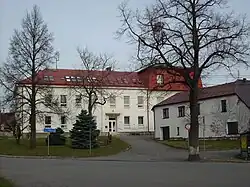 Bílá dvoupatrová budova s červenou střechou, před ní roste několik nižších stromů a keřů, vpravo roh další budovy