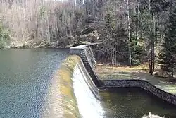 Pohled zblízka zboku na hráz přehrady Naděje s jejím přepadem valící se vody; nad hrází se zvedá terén se zimním opadaným nezasněženým lesem