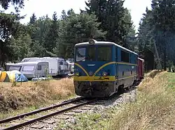 Modře zbarvená motorová lokomotiva v čele vlaku, jenž vede zalesněnou krajinou. V pozadí (na obrázku vlevo od lokomotivy) jsou stany a obytné přívěsné vozíky kempujících návštěvníků.