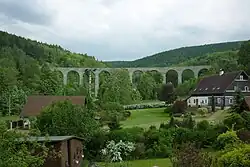 Kamenný víceobloukový viadukt přes údolí v obci Novina. V pozadí se nachází Ještědský hřbet (samotný nejvyšší vrchol však na tomto záběru není vidět). V blízkosti viaduktu jsou k vidění tradiční hrázděné domy a usedlosti. Snímek pochází z letního období a celá krajina je plná bující zeleně.