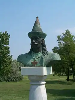 Obrázek tmavě zelenavé busty muže s kalpakem na hlavě, knírem a vlasy po ramena. Na krku muž má řetěz a na něm zavěšený kříž.