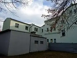 Fotografie zachycuje členitý zadní trakt budovy Záložny, který je opatřen novou zelenou omítkou. Popředí tvoří malý šedivý technický objekt a plot z vlnitého plechu.