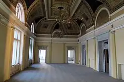 Fotografie zachycuje rozlehlý taneční sál v prvním poschodí Záložny. Sál je prázdný, parketová podlaha je léta neudržovaná a po topení zůstaly jen nosníky ve zdech. Strop sálu je bohatě zdoben štuky a freskami. Visí z něj dva lustry bez žárovek.