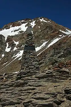 Fotografie kamenného pomníku v horách, na pozadí hora se zbytky sněhu