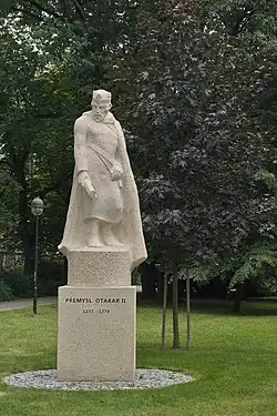 Fotografie kamenné sochy muže s pláštěm a korunou na hlavě stojící na podstavci v parku. V levé ruce drží svitek.