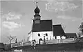 Černobílá fotografie kostela z meziválečné doby, kolem kostela jsou mladé stromky, v popředí stojí dvě děvčata