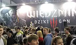 Procházející osoby na veletrhu před plakátem propagujícím hru