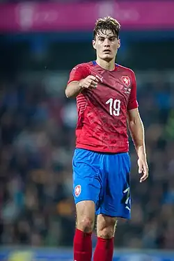 Fotbalista Patrik Schick v červeném dresu české reprezentace se dívá ukazuje pravou rukou směrem do objektivu.