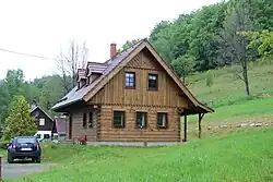 Dřevěný srub se šikmou střechou. Kolem domu se zelená louka. Před domem stojí automobil Dacia Duster