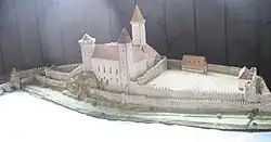 Fotografie modelu hradu, který má tři věže, stojí nad řekou a kolem něj jsou prostory ohraničené hradbami. Model se nachází ve vitríně.