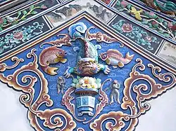 V centru fotografie hnědočervené plastické ornamenty na modré zdi, vlevo a vpravo nahoře pásy ornamentů. Motivy rostlin a ryb.