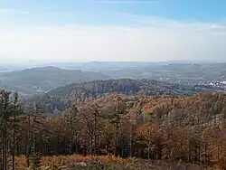V zelený kopec, okolní les žlutooranžově zbarvený, v pozadí tmavší kopce, nahoře modrá obloha s bílými mraky