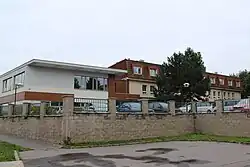 Zděné budovy stojící na zvýšeném dvorku za plotem. Na dvorku parkují osobní a dodávkové automobily. Budovy jsou dvoupodlažní, jen objekt vpravo má navíc ještě vestavěné podkroví. Stavba vlevo má rovnou střechu. Budova vlevo je omítnuta šedivě až světle modře, dům vpravo světle žlutě až okrově. Na dvoře před roste jehličnatý strom. Peřd plotem, na straně k pozorovateli, se nachází asfaltová plocha.