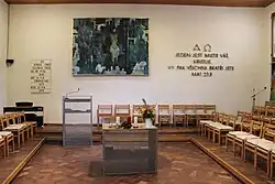 Pohled na bílou stěnu. Uprostřed visí rozměrný obraz laděný do převažujících modrých odstínů. Vpravo se nachází nápis „Jeden jest mistr váš, Kristus, vy pak všichni bratři jste“. Nalevo na zdi visí tabule s pořadem bohoslužby. Vpravo před ní se nachází kovová kazatelna. V přední části fotografie stojí kovový stůl Páně