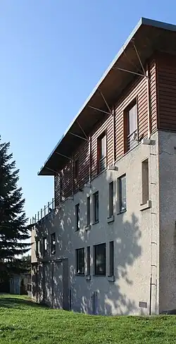 Pohled na stěnu objektu osvětlenou sluncem. Spodní dvě patra jsou zděná, horní je obložené dřevem. Ve zděné části se nacházejí obdélníková a čtvercová okna, ve vrchním patře okna francouzská. Objekt je zakončen vodorovnou střechou