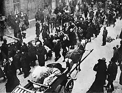 Ulice po níž kráčí dav lidí a vpředu proti směru chůze davu jede povoz tažený koněm.