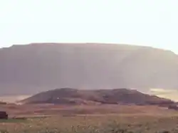 Fotografie se zeleným porostem ve předu a v pozadí s řekou a horami za ní. Uprostřed fotografie se nachází pozůstatek pyramidy