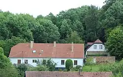 Dva zděné bíle omítnuté patrové domy se šikmou střechou, jeden k pozorovateli podélně, druhý čelem. Za nimi vzrostlý listnatý les, nad nímž je patrné světlé nebe. Před objektem podélného domu je ještě patrný hřeben střechy níže položené stavby.