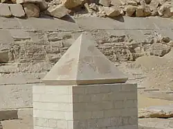 Pyramidion, který vypadá jako zmenšený model pyramidy, stojí na vyvýšeném kamenném stupínku. V pozadí se nachází zeď Červené pyramidy.