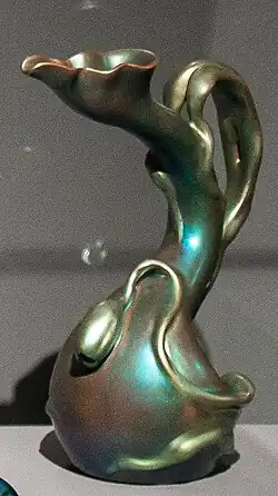 Secesní keramická nebo skleněná váza s organickým, tekoucím tvarem. Předmět má iridiscentní povrch s měňavými odlesky v zelených, modrých a bronzových odstínech. Tvar vázy je nepravidelný, vlnitý, s jedním výrazným uchem nebo výběžkem, který se elegantně stáčí. Horní okraj je zvlněný. Váza působí asymetricky a organicky a je vyfotografovaná na tmavém pozadí, což zvýrazňuje její lesklý povrch.