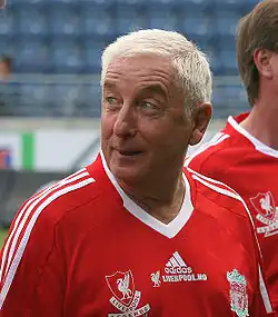 The head and upper torso of a gentleman in his late 50s. He has short white hair and is wearing a red football shirt, which has the Liverpool FC crest on the left breast, and a crest on the right breast that says "Liverpool Legends". A white logo of the Adidas sponsor is visible in the centre of the shirt, and three white stripes are present on the shoulder.
