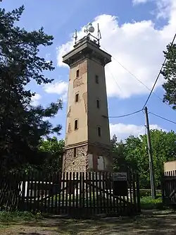 zděná oprýskaná věž, na vrcholu věže antény, v okolí vzrostlé stromy, na pozadí modré nebe s pár bílými oblaky