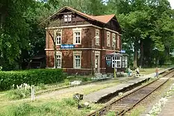 Nádraží v Rynolticích, představující typickou architekturu ATE s červenými cihlami, v letním období. Stanice je i v současnosti upravena a osázena množstvím okrasných květin; celá se pak nachází na okraji menšího listnatého lesa.