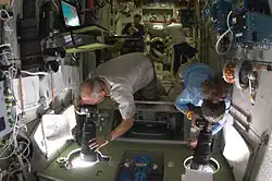 Astronauti NASA Jeffrey Williams a Nicole Stott z expedice 21 fotografují z okénka modulu Zvezda během přibližování lodi Atlantis, vlevo nahoře jsou vidět zavěšené další výměnné objektivy